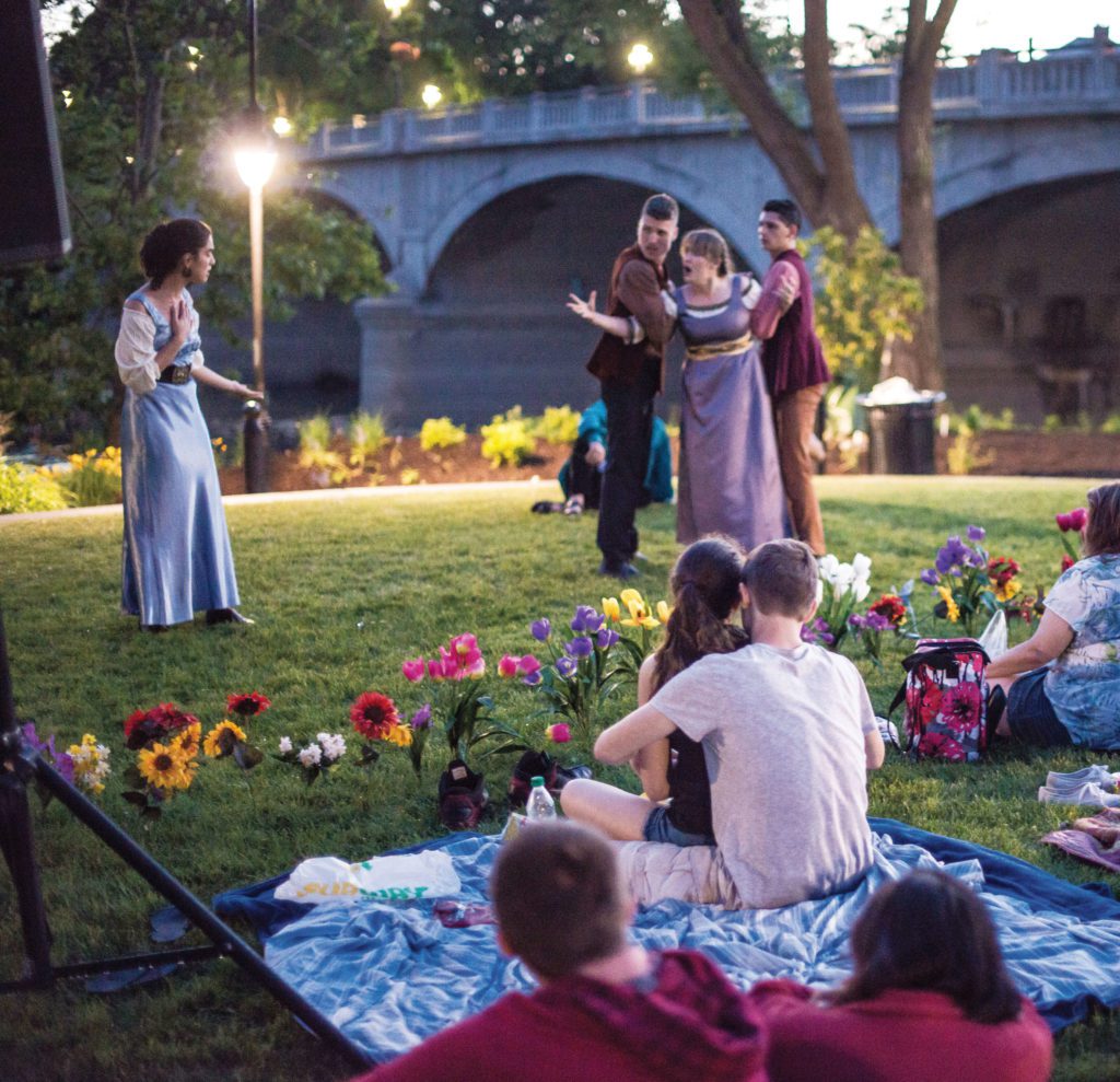 Actors put on Shakespeare in a park as audience watches from blankets.