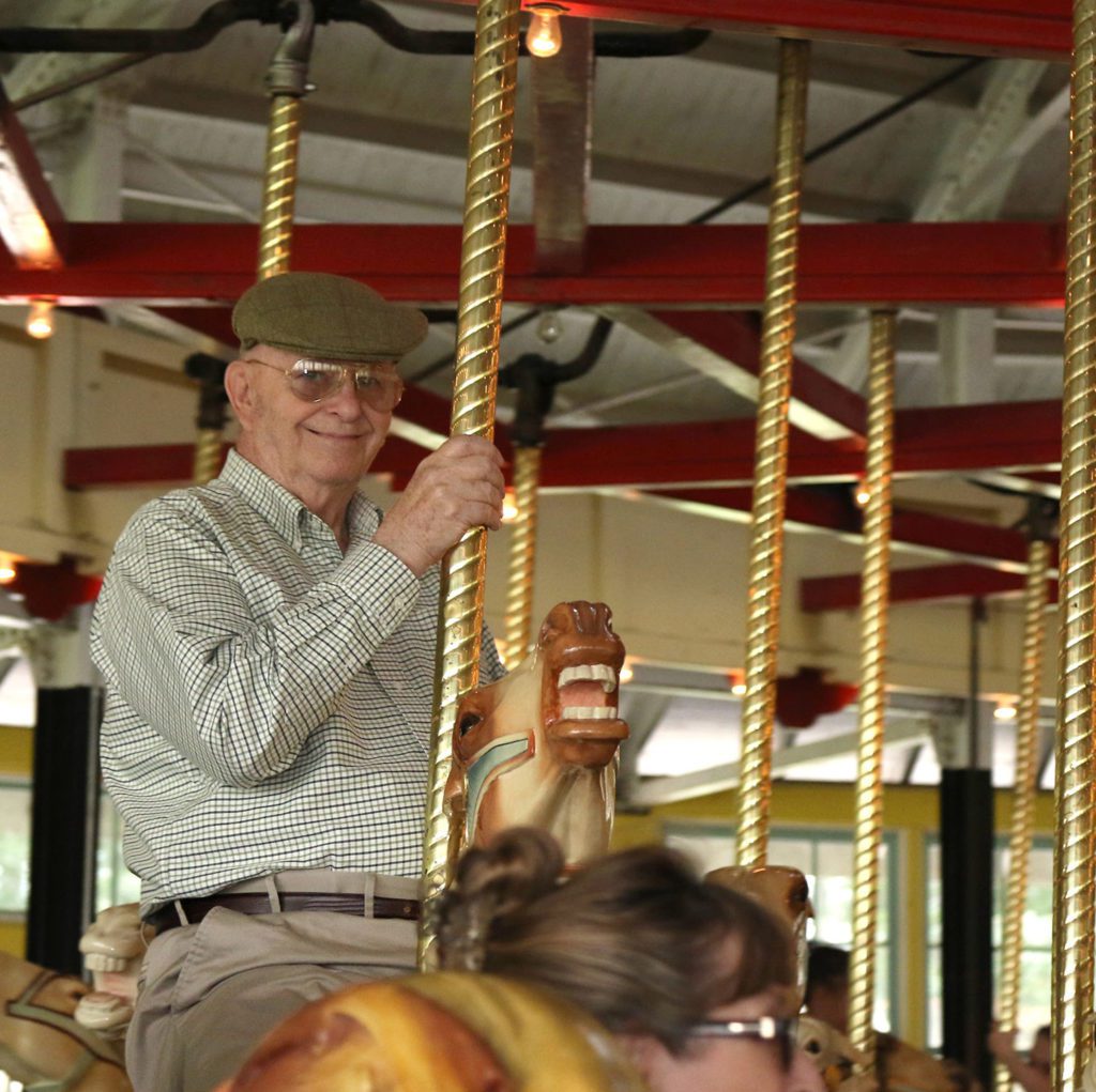 An elderly man smiling and riding a carousel horse.